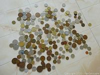 Photo of a large assortment of many foreign coins spread out on a light tile floor showing diversity in size, color and type.