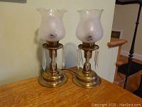 Two frosted glass buffet lamps with brass-tone bases and clear hanging glass prisms, placed on a wooden surface.