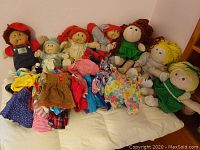 View of seven dolls sitting on a bed with multiple pieces of doll clothing laid out in front showing variety of styles and colors