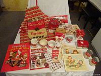 Assorted Campbell's Soup collectibles including apron, coloring book, recipe boxes, mugs, salt and pepper shakers, posters, magnet, cans, and five collectible Christmas ornaments in original packaging.
