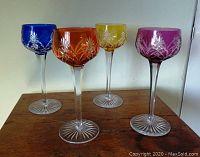 Four colored wine goblets in blue, orange, gold, and purple, shown on wooden surface with wall background.