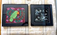 Two framed embroidered textile artworks displayed on a light patterned fabric, one with green parrot and pink flowers, the other with white floral and wave designs on dark background.
