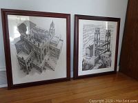Photo showing two framed black and white M. C. Escher poster prints leaning against a wall on a wooden floor.
