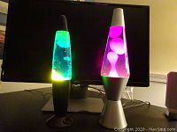 Two lava lamps side by side on a desk in front of a computer monitor; the left lamp is green and yellow with black base, the right lamp is pink and purple with silver base.