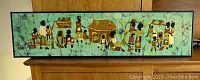 Framed fabric or canvas artwork depicting an African village scene with multiple figures and buildings on a green fabric background in black frame. Size approximately 35" x 9".