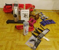 Mini Trainer pedal exercise machine with digital display, pink dumbbells, wooden dumbbell, hand grips, yoga straps, spring collars, ankle weights, cooling towel, and red gym bag on parquet floor.