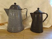 Two vintage enamelware pitchers with spatterware pattern shown upright, grey with dark speckled design, one larger and one smaller