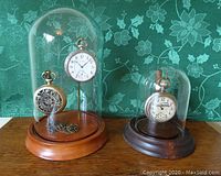 Front view of two dome display cases on wood bases with pocket watches under glass domes, one with a lighter wood base and one with a darker wood base.