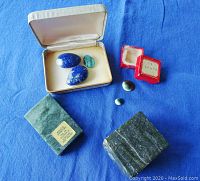 Photo of various polished stones including lapis lazuli and turquoise in display boxes, greenish Nephrite blocks on blue cloth background