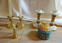 Two pairs of candlesticks with a brass candle snuffer and a ceramic box with sun motif, displayed on a wooden floor.
