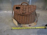 Front view of vintage wicker fishing creel showing hinged lid, rope handle, shoulder strap, and measuring tape indicating approximate width of 12 inches.