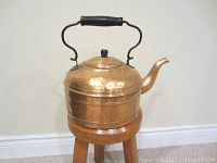 Antique copper kettle shown from an angle on a wooden stand against a plain white wall. The kettle has a curved spout, black handle, and a round lid with knob. Surface shows wear and patina.