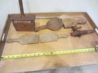 Photo shows seven vintage wooden household utensils arranged on wooden tray including butter mold, paddles, and crank handle. A tape measure below shows scale.