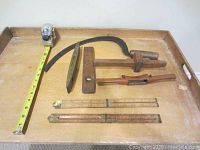 Photo showing six vintage woodworking tools on a wooden tray: woodworking scribe, shave, two small spirit levels, two folding rulers, hand scythe.