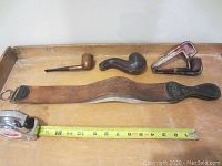 Photo showing three vintage tobacco pipes and a leather barber strop on a wooden surface with measuring tape for scale.