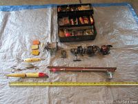 Photo showing four fishing reels, two fishing rods, a plastic tackle box with assorted lures and fishing accessories spread out on a tarp with a measuring tape for scale.