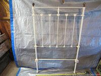 Single iron headboard painted white with decorative spindles and floral motifs, spherical top finials, set against blue tarp.