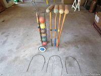 Photo showing 4 wooden croquet mallets, 6 multi-colored balls in a metal cart with blue wheels, and 3 metal wire hoops on ground.