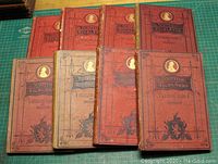 Set of eight hardcover books with red and ochre cloth covers and gilt titles and decoration on spines.