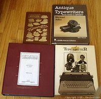 Four books laid out on wooden floor: three hardcover books about antique typewriters and one red binder with repair instructions.