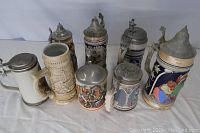 Eight vintage German beer steins with pewter lids in used condition, arranged from smallest to tallest.