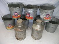 Seven galvanized milking pails lined in two rows and two galvanized cream cans with wooden lids in front, placed on a white cloth background.