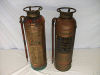 Two vintage copper fire extinguishers side by side with attached hoses and handles