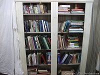 Full front view of two-door white bookcase filled with various hardcover and softcover books on all five shelves, showing large collection with mixture of fiction and nonfiction titles.