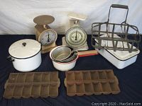 Full view of milk bottle carrier, two cast iron muffin pans, two vintage country scales, and enamelware pots arranged on a dark cloth.