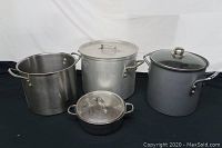 Four large stock pots with lids of varying sizes and materials on black cloth, includes stainless steel, aluminum, and black aluminum with glass lid