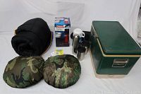 Photo showing camping gear including black rolled nylon sleeping bag, two camo bun warmers, green Coleman cooler, thermos and coffee warmers, and boxed new Coleman LED lantern.
