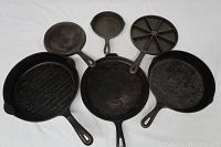 Six cast iron pans displayed on white cloth, showing variety of pan types, handles, and skillet shapes.