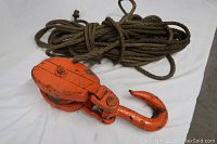 Orange metal block and tackle pulley with hook and thick gauge brown rope coiled behind it.