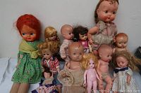 Grouping of 14 dolls with varied clothing and condition, arranged on table against wall