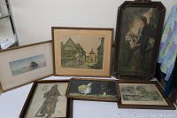 Six framed artworks and prints laid on table and leaning against wall showing overall lot condition.