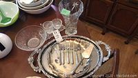 Photo of silver trays and assorted silver plate utensils including collectible spoons on a table with other glass pieces