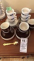 Two sets of espresso cups stacked on white saucers with text on cups. Also shows four dark brown espresso cups stacked with matching saucers. A set of six ornate gold-tone spoons are displayed in front.
