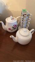 Two ceramic teapots on a wooden surface: one with rose floral design and one plain white textured teapot