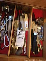 Top view of drawer showing multiple flatware pieces including forks, spoons, steak knives, pizza cutter, tongs, scissors, and a souvenir bottle opener.