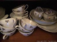 Stack of tea cups and saucers alongside creamer and sugar bowl with lid, all in white china with gold floral decoration.