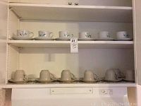Photo showing 7 cups and 7 saucers of 'The Classics' pattern on top shelf and 6 cups and 6 saucers of 'Fascino' pattern on bottom shelf, displayed in shelving unit.