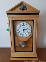 Front view of wooden mantle clock with white dial, Roman numerals, and visible pendulum.