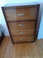 Front view of mid-century wooden chest of drawers with four drawers and metal wing-shaped handles.