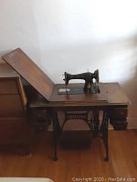 Front angled view of vintage Singer sewing machine on wooden treadle table with wrought iron frame and foot pedal.