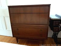 Front view of mid-century modern wood tallboy chest showing 3 drawers with integrated handles and tapered legs on casters.