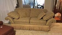Full front view of champagne colored upholstered sofa with cushions and fringe skirt.