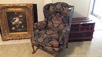 Wingback chair with floral pattern upholstery near painting and red chest, showing front and side views.