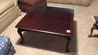 Front-side angle view of the square wooden coffee table showing the glossy finish, inlaid border, and cabriole legs on a carpeted floor.