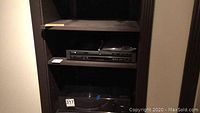DVD player and accessories on a dark wooden shelf, showing the Toshiba DVD player, remote control on top, and the owner's manual.