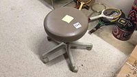 Single cushion stool with round brown padded seat and gray plastic 5-leg base with wheels, shown from different angles on carpeted floor near tennis rackets and other items.